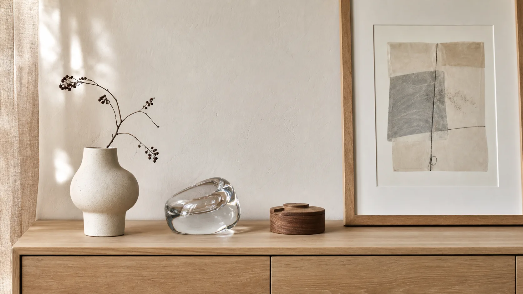 Keramisk vas, klart glasobjekt och litet träobjekt på ett ljust sideboard bredvid en tavla